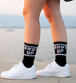 Qorapa LaraLara black crew socks with white SHUT UP text and striped cuffs worn with white sneakers outdoors
