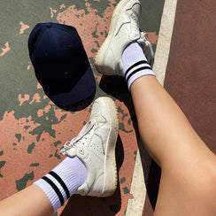 Qorapa LaraLara white low top court sneakers with white striped socks on a tennis court next to a navy baseball cap