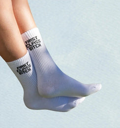 Qorapa LaraLara white crew socks with black First Class Bitch graphic text on ribbed cuffs worn on feet