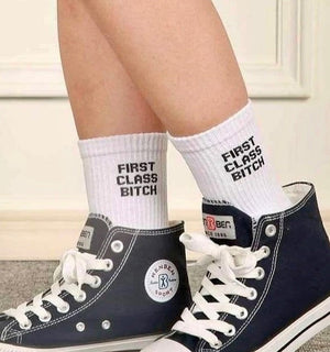 Qorapa LaraLara white crew socks with black First Class Bitch graphic text worn with black high-top sneakers