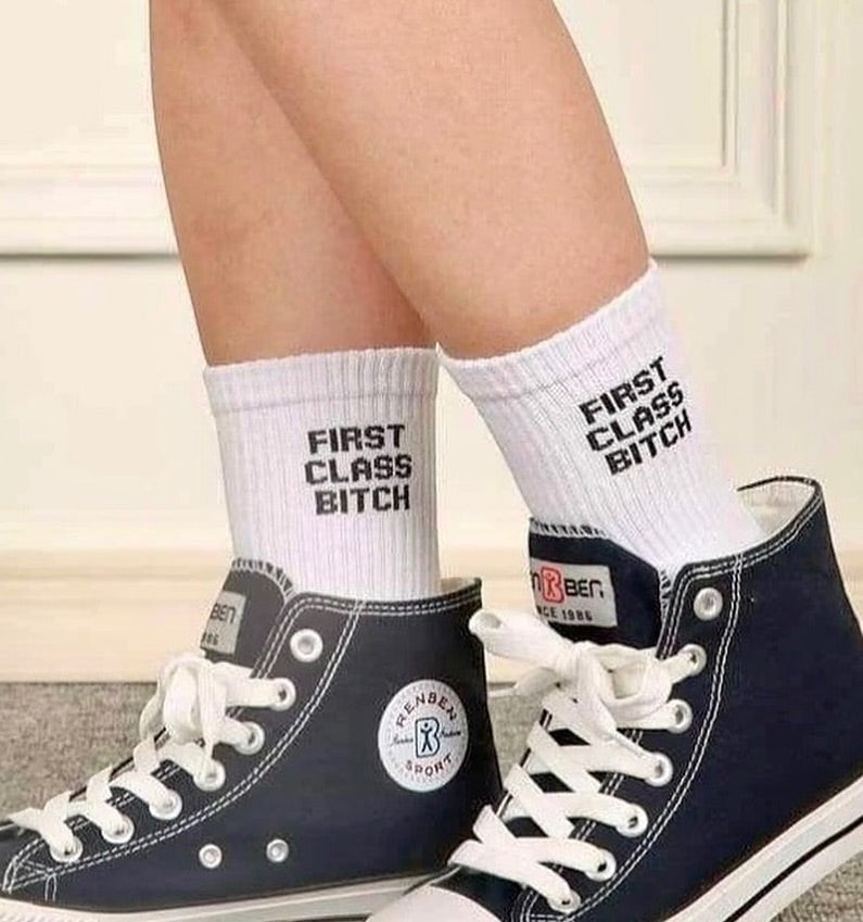 Qorapa LaraLara white crew socks with black First Class Bitch graphic text worn with black high-top sneakers