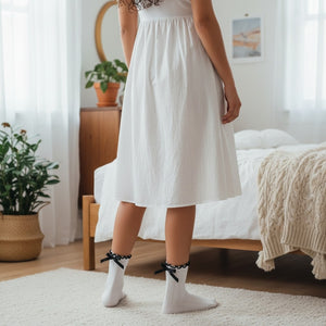 Qorapa LaraLara white ankle socks with ruffled trim and black ribbon bows worn with a white dress in a bedroom.