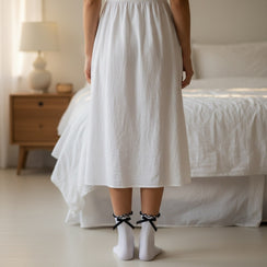 Qorapa LaraLara white ankle socks with ruffled cuffs and black ribbon bows worn with a white skirt in a bedroom