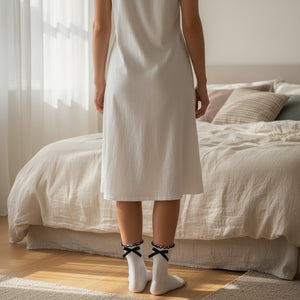 Qorapa LaraLara white ankle socks with ruffled cuffs and black ribbon bows worn by a woman in a white nightgown.